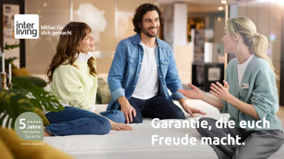Ein Paar sitzt auf einem Bett und unterhält sich mit einem Verkäufer in einem Interliving-Möbelhaus. Dabei wird auf die 5-jährige Garantie hingewiesen.