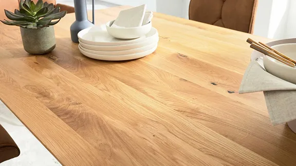 Nahaufnahme einer hellen Massivholz-Tischplatte mit natürlicher Maserung, Geschirr und einer kleinen Pflanze.